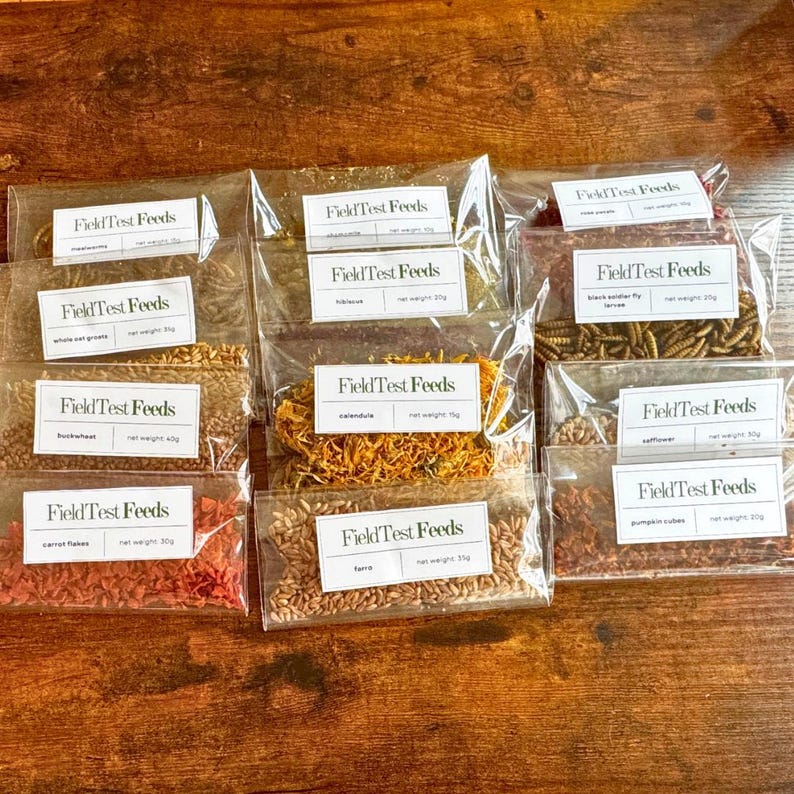 Puede incluir: Una colecci&oacute;n de bolsas de pl&aacute;stico transparente llenas de varios ingredientes secos, cada una etiquetada con "Field Test Feeds". Los ingredientes incluyen granos de avena entera, trigo sarraceno, copos de zanahoria y cal&eacute;ndula. Las bolsas est&aacute;n dispuestas sobre una superficie de madera.