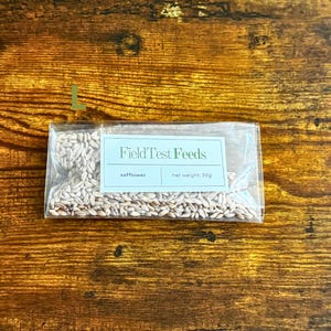 Puede incluir: Una bolsa de pl&aacute;stico transparente llena de semillas de c&aacute;rtamo, con una etiqueta que dice "Field Test Feeds". La etiqueta tambi&eacute;n indica un peso neto de 30g. La bolsa est&aacute; sobre un fondo de madera.