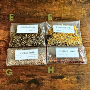 Puede incluir: Cuatro bolsas de pl&aacute;stico transparente de productos FieldTest Feeds se muestran sobre una superficie de madera. Las bolsas contienen larvas de mosca soldado negra, cal&eacute;ndula, farro y cubos de calabaza. Cada bolsa tiene una etiqueta blanca.