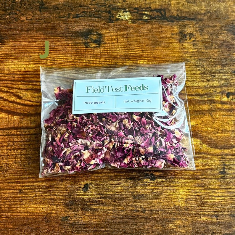 Puede incluir: Una bolsa de pl&aacute;stico transparente llena de p&eacute;talos de rosa secos. La etiqueta dice "FieldTest Feeds", "rose petals" y "net weight: 10g". Los p&eacute;talos son una mezcla de colores rosa y burdeos. El fondo es una superficie de madera.