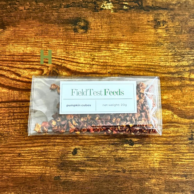 Puede incluir: Un paquete transparente con cubos de calabaza, con el texto "Field Test Feeds" y "pumpkin cubes" impreso en una etiqueta blanca. El peso neto es de 20g. El paquete est&aacute; sobre una superficie de madera.