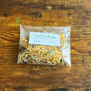 Puede incluir: Una bolsa de pl&aacute;stico transparente llena de flores de cal&eacute;ndula secas, etiquetada como "Field Test Feeds". La etiqueta tambi&eacute;n indica un peso neto de 15g. La bolsa est&aacute; sobre una superficie de madera.