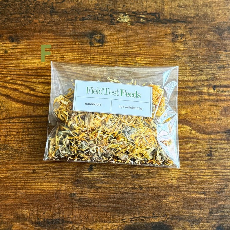 Puede incluir: Una bolsa de pl&aacute;stico transparente llena de flores de cal&eacute;ndula secas, etiquetada como "Field Test Feeds". La etiqueta tambi&eacute;n indica un peso neto de 15g. La bolsa est&aacute; sobre una superficie de madera.