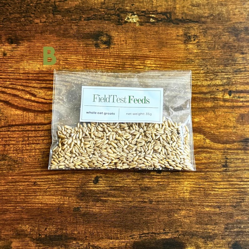 Puede incluir: Una bolsa de pl&aacute;stico transparente llena de granos de avena enteros, con una etiqueta que dice "FieldTest Feeds". La etiqueta tambi&eacute;n indica "whole oat groats" y "peso neto: 35g". La bolsa est&aacute; sobre una superficie de madera.