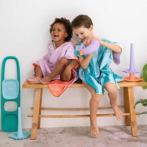 May include: Two children wearing striped hooded towels sit on a wooden bench. One child wears a purple and white striped towel with an orange trim, while the other wears a blue and green striped towel. A teal bath toy and a stacking toy are nearby.