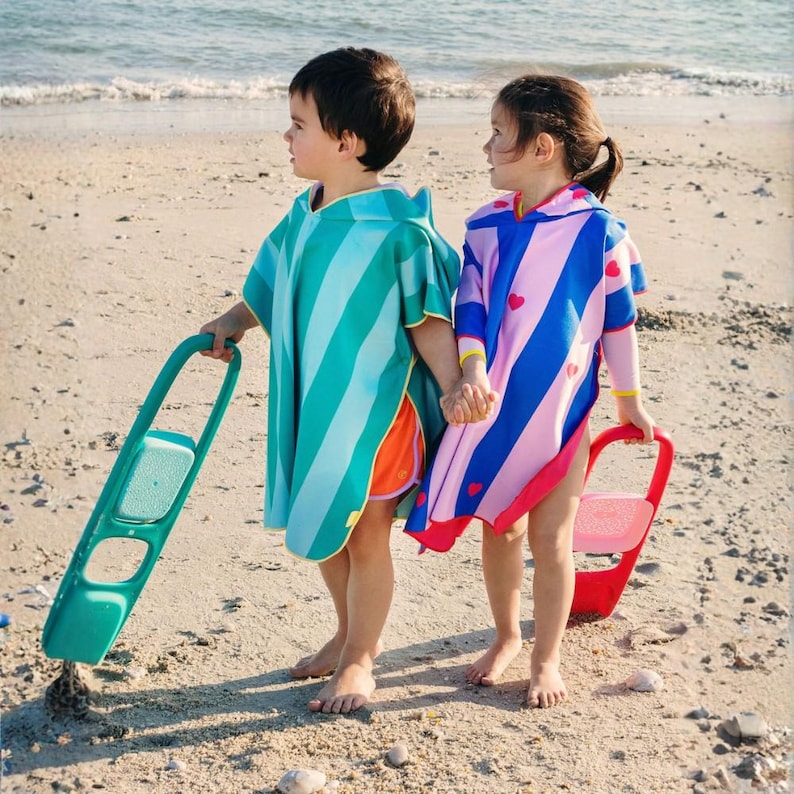 May include: Two children on a sandy beach wearing colorful striped hooded beach ponchos. One poncho is teal and blue, the other is pink and blue with heart accents. Each child holds a beach toy.