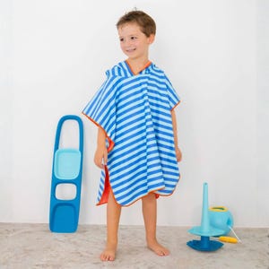 May include: A child wearing a blue and light blue striped hooded beach towel with orange trim. The towel is a poncho style. A blue and light blue toy is on the floor to the right. A blue toy with a handle is on the left.