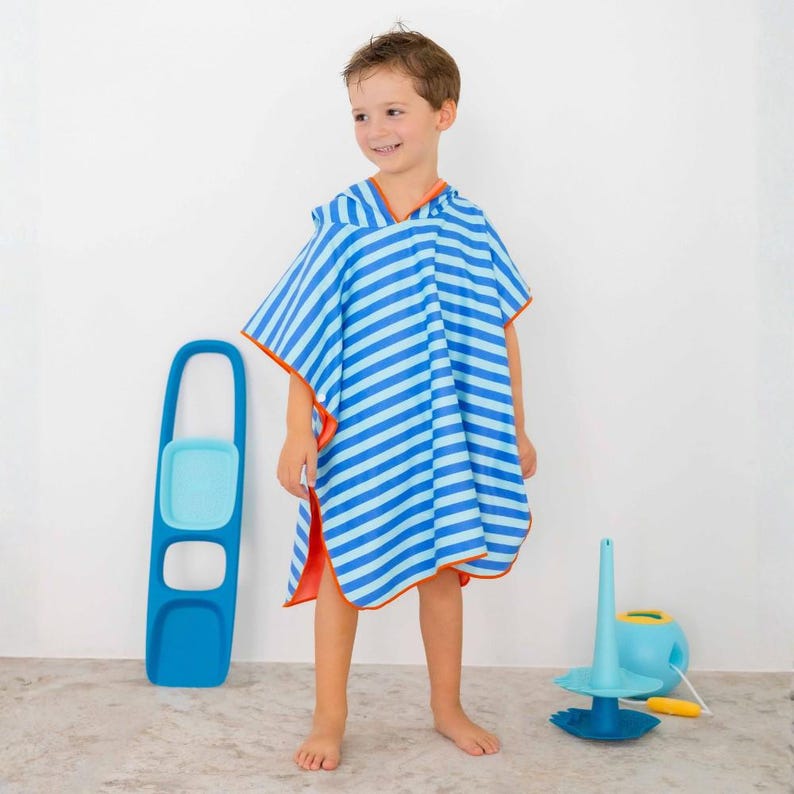 May include: A child wearing a blue and light blue striped hooded beach towel with orange trim. The towel is a poncho style. A blue and light blue toy is on the floor to the right. A blue toy with a handle is on the left.