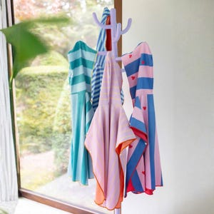 May include: A lavender coat rack holds several colorful hooded towels. The towels feature stripes and heart patterns in shades of blue, pink, and orange. The towels are hanging in front of a window.