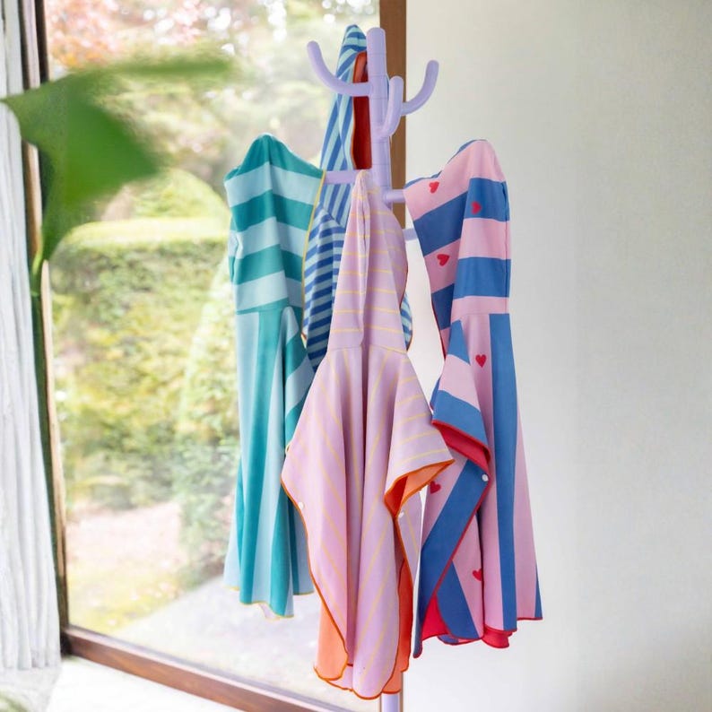 May include: A lavender coat rack holds several colorful hooded towels. The towels feature stripes and heart patterns in shades of blue, pink, and orange. The towels are hanging in front of a window.