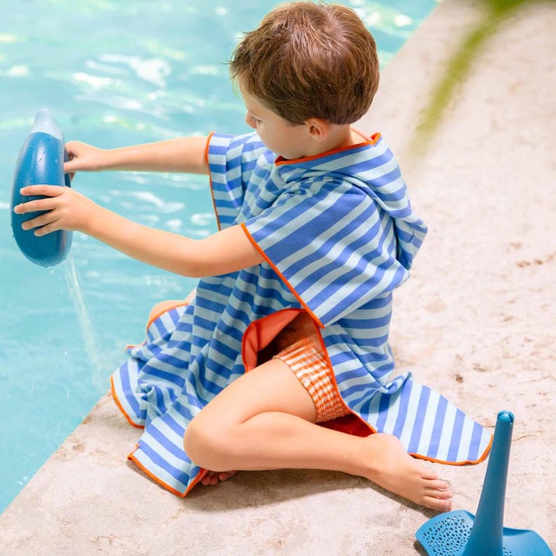 May include: A blue and white striped hooded beach towel with orange trim. The towel is wrapped around a person sitting near a pool. The person is also wearing orange and white striped swim trunks. A blue water bottle is being held.