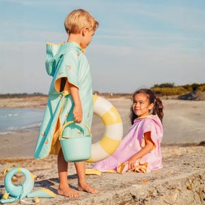 May include: Two children on a beach wearing hooded beach towels. One child wears a light blue towel with sailboat designs and holds a matching bucket. The other child wears a pink towel with white polka dots. A yellow and white inflatable ring is nearby.
