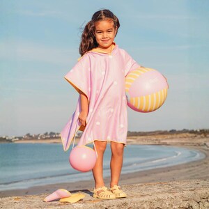 May include: A young person stands on a stone wall at the beach, wearing a pink polka-dot hooded beach towel with yellow trim. They hold a pink and yellow striped beach ball and a pink balloon. Yellow sandals and a yellow sun hat are at their feet.