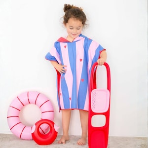 May include: A child wearing a blue, pink, and red striped hooded beach poncho with red heart accents. A pink inflatable ring and a red water toy are on the floor. A red beach toy is held by the child.