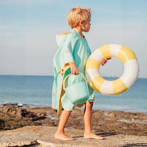 May include: A child wearing a light blue beach poncho with a yellow hood and watermelon graphics, holding a matching bucket and a yellow and white striped inflatable ring. The scene is set on a rocky beach with the ocean in the background.