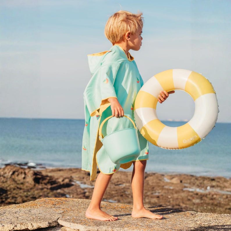 May include: A child wearing a light blue beach poncho with a yellow hood and watermelon graphics, holding a matching bucket and a yellow and white striped inflatable ring. The scene is set on a rocky beach with the ocean in the background.