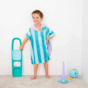 May include: A child wearing a blue and turquoise striped hooded beach towel. The towel has a lavender hood and yellow trim. A teal-colored toy is held in one hand. Other toys are on the floor, including a purple and blue toy and a blue ball.