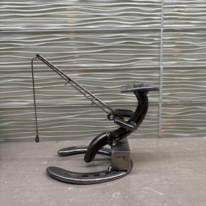 May include: A metal sculpture of a person fishing, crafted from horseshoes and metal rods. The figure is seated, holding a fishing rod with a line and hook. The sculpture is a unique decorative item.