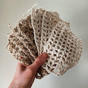 Puede incluir: Una mano sostiene una colección de bolsas tejidas, de color beige y bronceado, con forma de red. Las bolsas están hechas de fibras naturales, con una variedad de texturas y tejidos. Algunas bolsas tienen un patrón más abierto, similar a una rejilla, mientras que otras están tejidas apretadamente.