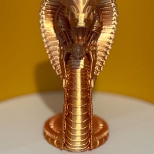 May include: A gold-coloured cobra sculpture with detailed mechanical elements. The cobra is depicted upright, with its hood flared and an engine design integrated into the head. The sculpture is on a white surface with a yellow backdrop.