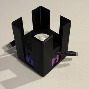 May include: A black, cube-shaped electronic device with a white interior and multiple ports. Cables extend from the sides. The device has a modern design and appears to be a charging station or hub.