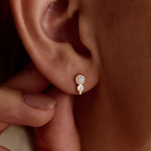 May include: Gold stud earrings with an opal and a clear gemstone. The opal is round and above a smaller, round, clear gemstone. The earrings are shown on an ear, highlighting their delicate design and elegant simplicity.