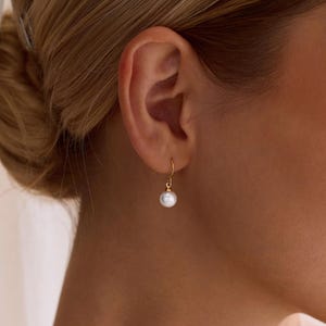 May include: A gold-colored, small hoop earring with a single, white pearl. The earring is worn on the ear of a person with blonde hair. The earring is a simple, elegant design.