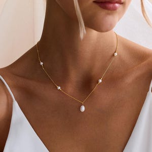 Könnte beinhalten: Eine zarte goldene Halskette mit kleinen, runden Perlen und einem einzelnen, tropfenförmigen Perlenanhänger. Die Halskette wird zu einem weißen Kleid getragen, was auf Braut- oder formelle Anlässe hindeutet.