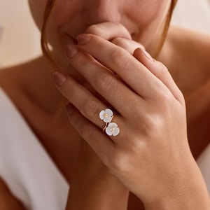 May include: A gold-toned ring featuring two white flower-shaped elements, each with a small central detail. The ring is worn on a finger, showcasing the delicate design of the two petals arranged in a bypass style on a thin metallic band.