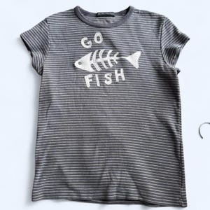 Op de afbeelding: Grijs en wit gestreept T-shirt met de tekst "GO FISH" en een visgraatmotief. Het T-shirt heeft korte mouwen en een ronde hals. De tekst en de afbeelding zijn wit.