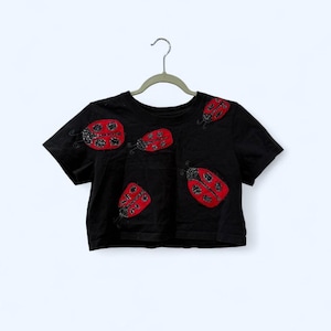 Peut inclure: Haut court noir avec des appliqués coccinelles rouges. Les coccinelles ont des détails perlés noirs et sont disposées sur le devant du t-shirt. Le t-shirt est suspendu à un cintre gris sur fond blanc.