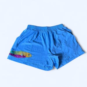 Peut inclure: Short bleu clair avec une taille élastique et une application de poisson coloré sur la jambe gauche. Le motif de poisson présente des nuances de jaune, vert, violet et rose. Le short est en tissu doux et léger.