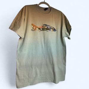 Peut inclure: T-shirt à manches courtes avec un dégradé de couleurs, allant du beige en haut au bleu-vert en bas. Le t-shirt présente une application de poisson avec des motifs orange, dorés et bleus.