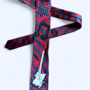 Może przedstawiać: Czerwono-niebieska w paski krawat z haftowanymi patriotycznymi i muzycznymi wzorami. Krawat posiada gwiazdę, nutę muzyczną i gitarę. Gitara jest srebrna i biała z czerwonym gryfem.