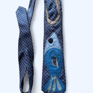 Peut inclure: Une cravate bleu marine avec un motif de losanges répétés. La cravate présente un grand motif de poisson bleu appliqué avec un œil noir et un accent gris. Le poisson est fait de différentes nuances de tissu bleu. Un accessoire unique.