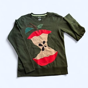 May include: An olive green sweatshirt with a large applique of a partially eaten apple. The apple design includes red, tan, and black details, with green and brown leaf accents. The long-sleeved top has a crew neck.