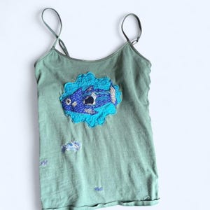 Op de afbeelding: Saliegroene camisole met dunne bandjes. De top heeft een grote blauwe visapplicatie in het midden, omgeven door een turquoise wolkvorm. Extra kleinere visapplicaties zijn aanwezig.