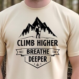 May include: A beige t-shirt with the text "CLIMB HIGHER BREATHE DEEPER" in black. The design includes a mountain range silhouette with a hiker and trees. The shirt is short-sleeved and made of a soft material.