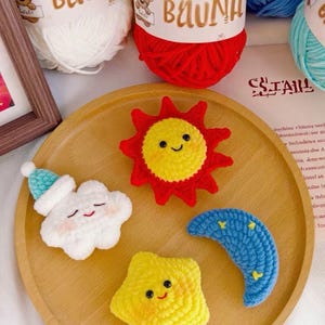 May include: A wooden tray holds four crocheted celestial shapes: a smiling sun, a star, a crescent moon, and a cloud with a hat. The sun is red and yellow, the star is yellow, and the moon is blue. White yarn balls with the text "BAUNH" are in the background.