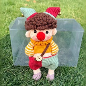 May include: A handmade crochet doll dressed as a jester, featuring a red nose, a yellow and white striped shirt, and red and green shorts. The doll has a brown, textured hair and a red and green hat with bells. It is inside a clear plastic box.
