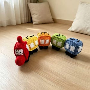 May include: A crochet toy train set consisting of five connected cars in red, yellow, orange, green, and blue. Each car features black wheels and white square windows stitched onto the sides. The red engine car is at the front of the line.