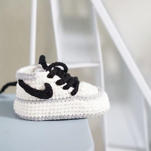 May include: A crochet baby shoe featuring a white base with light gray trim and a black crocheted swoosh logo on the side. The shoe includes thick black yarn laces tied in a bow at the top, with a high-top silhouette and a textured yarn construction.
