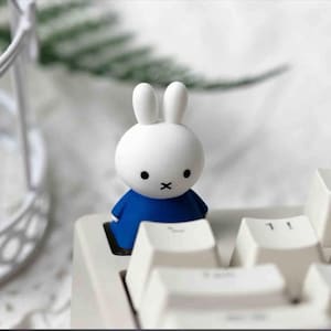 Peut inclure: Une figurine de lapin blanc avec des yeux noirs et une bouche en forme de 'X' noir, vêtue d'une robe bleue. La figurine est placée sur un clavier blanc, avec les touches visibles au premier plan. L'arrière-plan est flou.