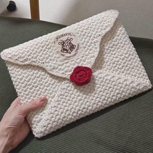 May include: An off-white crocheted envelope-style pouch featuring a textured stitch pattern. A rectangular fabric patch with the Hogwarts crest and text is sewn on the flap, secured by a circular, dark red crocheted wax seal-style button.