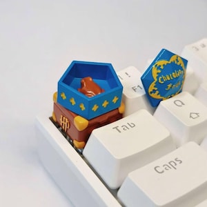 May include: A keyboard with custom keycaps featuring a brown chest, a blue hexagonal box with a frog, and a blue chocolate bar. The keycaps are placed on a white keyboard.