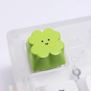 May include: A bright lime green mechanical keyboard keycap shaped like a four-leaf clover. The top surface of the clover features a simple, minimalist black printed face with two small circular eyes and a small v-shaped mouth centered on the petals.