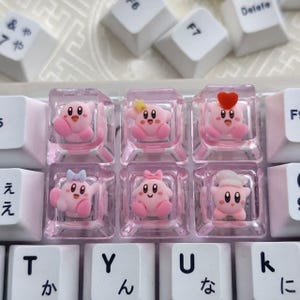 May include: Six clear mechanical keyboard keycaps featuring small, pink, round character figures. The figures include variations with a yellow star, a red heart, a blue bow, a pink bow, and a white chef hat, all set against a white keyboard.
