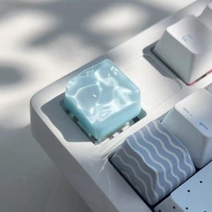 May include: A close-up of a white keyboard with custom keycaps. One keycap is a translucent, light blue square with a water ripple effect. Other keycaps are white and blue with wave patterns. The keyboard is in natural light.