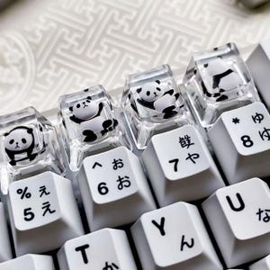May include: Close-up of a white keyboard with custom keycaps. The keycaps feature clear, transparent tops with small, black and white panda bear figures inside. The keyboard keys have Japanese characters and numbers.