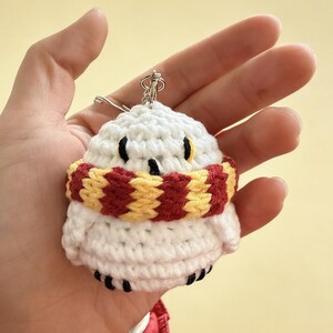 May include: A white crochet owl keychain with black eyes and feet, wearing a red and yellow striped scarf. The owl is held in a hand, showcasing the handmade details and the silver keychain ring.